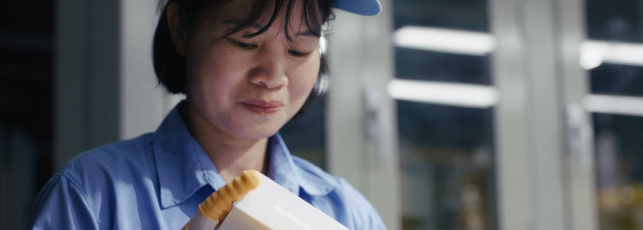 Funcionária com uniforme de trabalho de corpo inteiro e óculos de proteção observa a montagem do Apple Watch