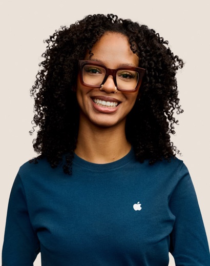 길고 곱슬곱슬한 검은 머리에 안경을 쓴 채 미소 짓고 있는 Apple Retail 팀원의 인물 사진.