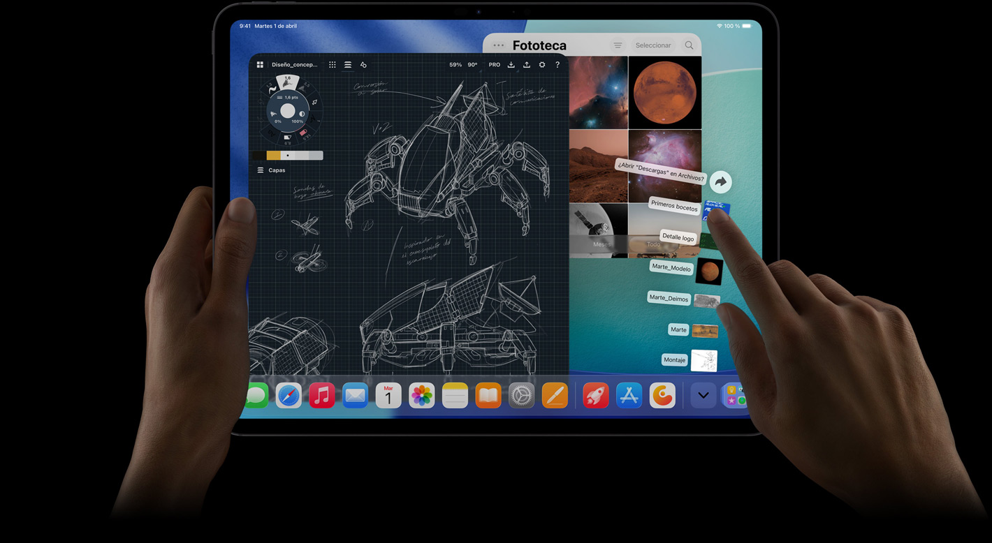 iPad Pro, parte delantera exterior, posición horizontal, la mano derecha hace un gesto sobre la carpeta Descargas con los archivos desplegados en abanico, la pantalla muestra la interfaz de iPadOS 26 con varias apps abiertas, entre ellas una app de edición de fotos con planos de robots, anotaciones manuscritas y varias etapas de diseño, y la Fototeca