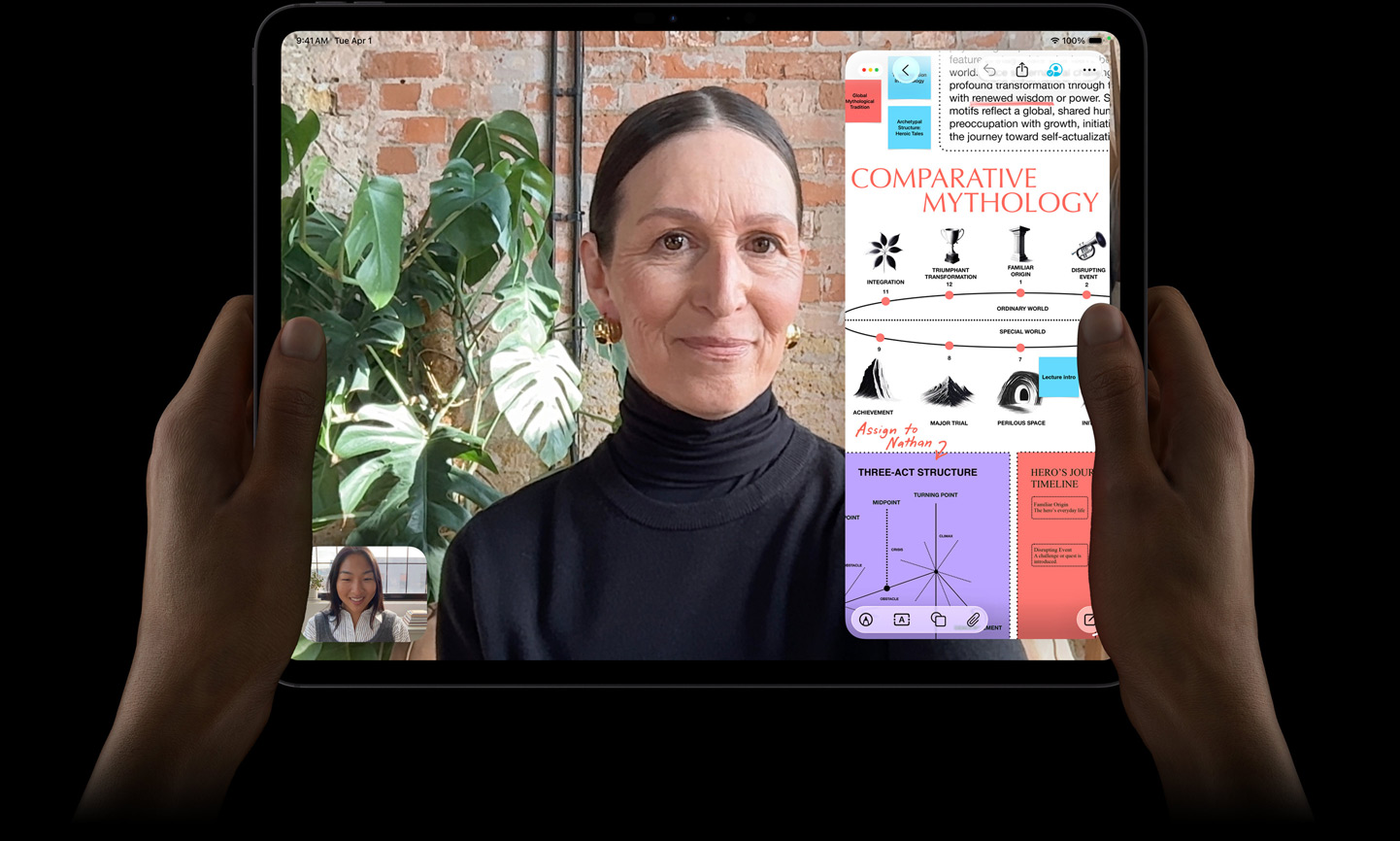 iPad Pro, face avant, noir sidéral, mains gauche et droite tenant l’appareil en orientation paysage, angles arrondis, cadre d’écran noir, écran affichant une conversation FaceTime traduite en direct et une app éducative montrant une présentation de la mythologie comparée avec du texte, des diagrammes et des notes manuscrites