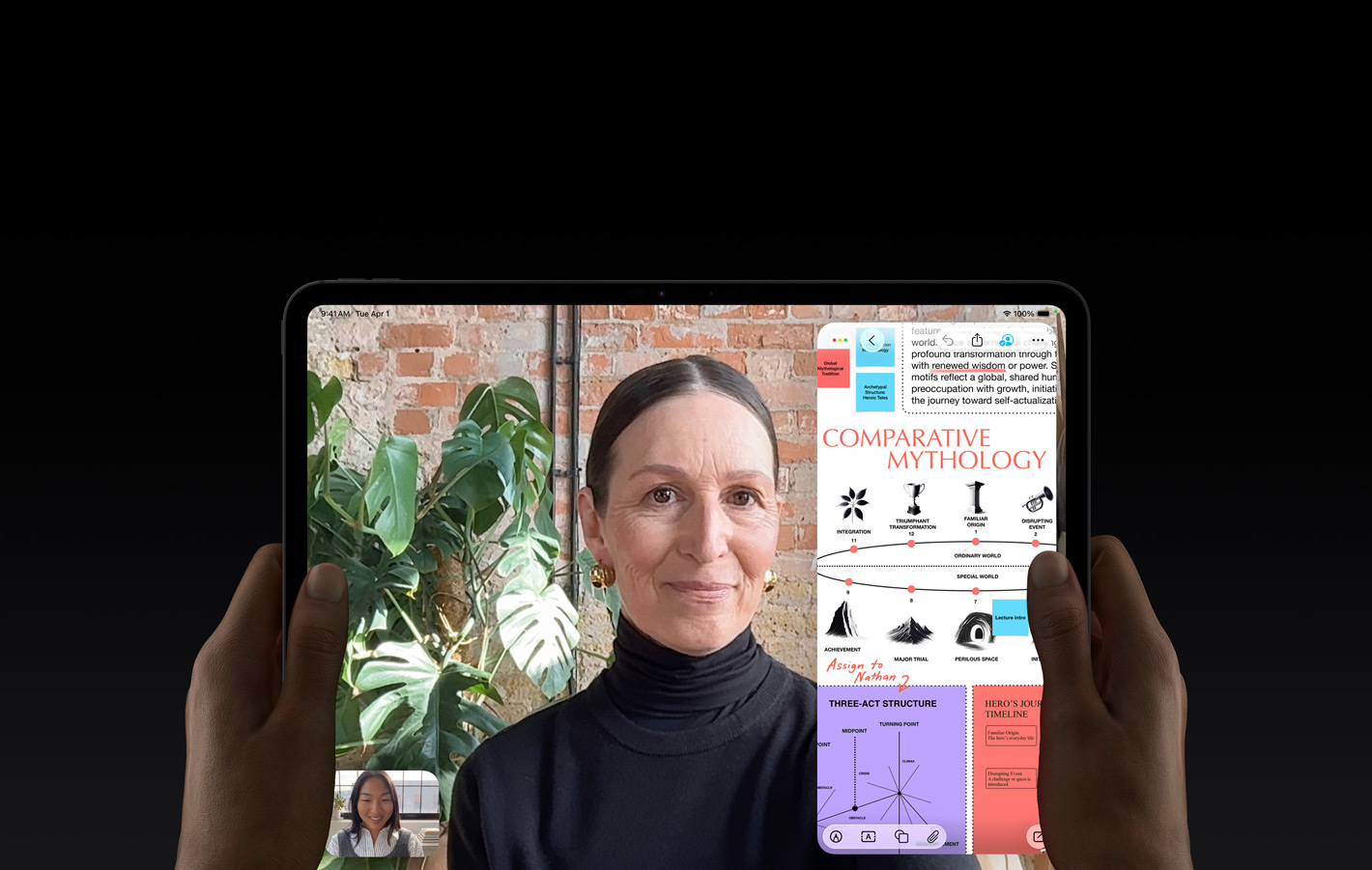 iPad Pro i space black vist forfra, et par hænder holder enheden i vandret position, afrundede hjørner, sort skærmkant; skærmen viser en FaceTime-samtale, der oversættes live, og en uddannelsesapp, der viser en præsentation af “Comparative Mythology” med tekst, diagrammer og håndskrevne noter