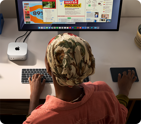 شخص يطبع وهو جالس إلى مكتب مع جهاز Mac mini، شاشة Studio Display متصلة، لوحة مفاتيح ماجيك، لوحة تعقب ماجيك، أغراض متنوعة