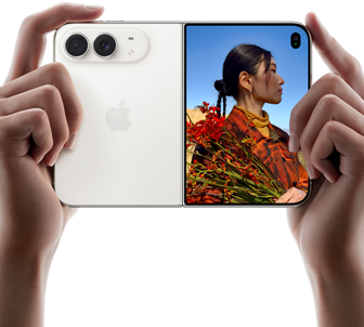 iPhone Duo, culoare Alb stelar, ținut de două mâini în dispunere orizontală, desfășurat, partea din stânga prezintă camerele spate Dual Fusion de 48 MP, partea din dreapta prezintă o persoană care se pozează pe ecranul extern, subiectul pozelor poate vedea chipul său în timp ce poza este făcută cu ajutorul aplicației Previzualizare Duo
