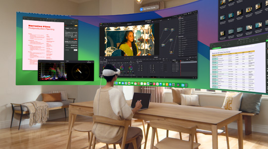A person wearing Apple Vision Pro sits at a desk with a 13-inch MacBook Pro open, and a curved, ultra-wide display of their screen contents projects into the room