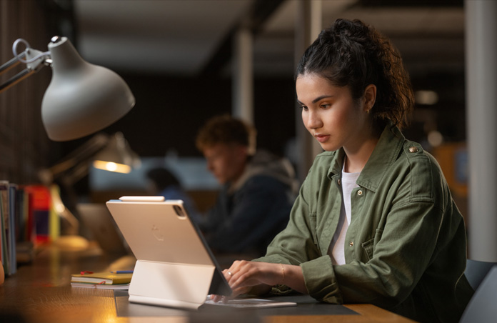Une étudiante assise à un bureau dans un espace de travail tape sur un iPad avec un Magic Keyboard, illustrant la polyvalence des appareils Apple