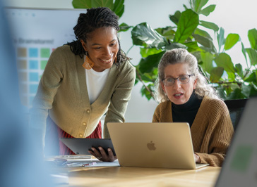 Deux professeures collaborent à un bureau; l’une tient un iPad et l’autre travaille sur Mac