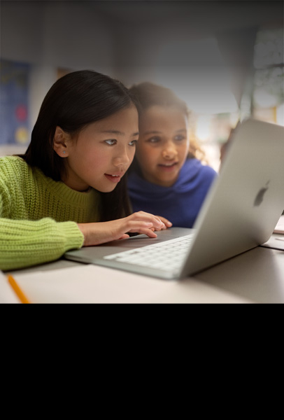 Two young students seated at a desk looking intently at a MacBook as they collaborate in a classroom
