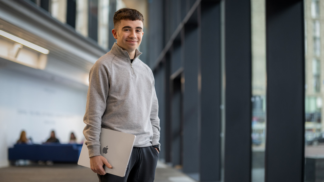 Un estudiante de pie en el pasillo de un edificio moderno con un portátil MacBook, sonriendo a la cámara