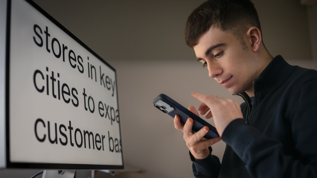 Un estudiante utiliza un iPhone sentado frente a un monitor que muestra el contenido de su clase de empresariales con el texto ampliado