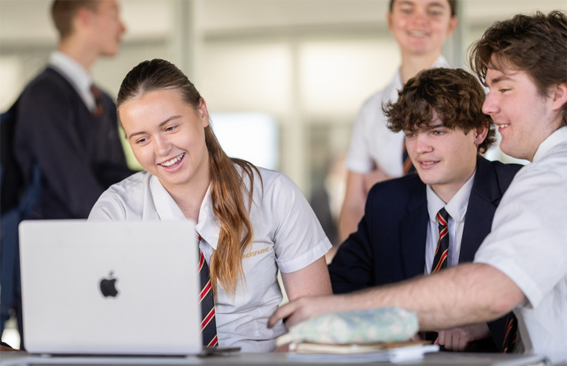 Un grupo de estudiantes con uniformes escolares se congrega alrededor de un MacBook, sonríen y trabajan en equipo.