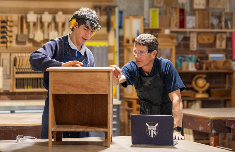 Un profesor de carpintería y un estudiante inspeccionan un pequeño mueble de madera. Hay un portátil de la escuela en la mesa de trabajo delante de ellos.