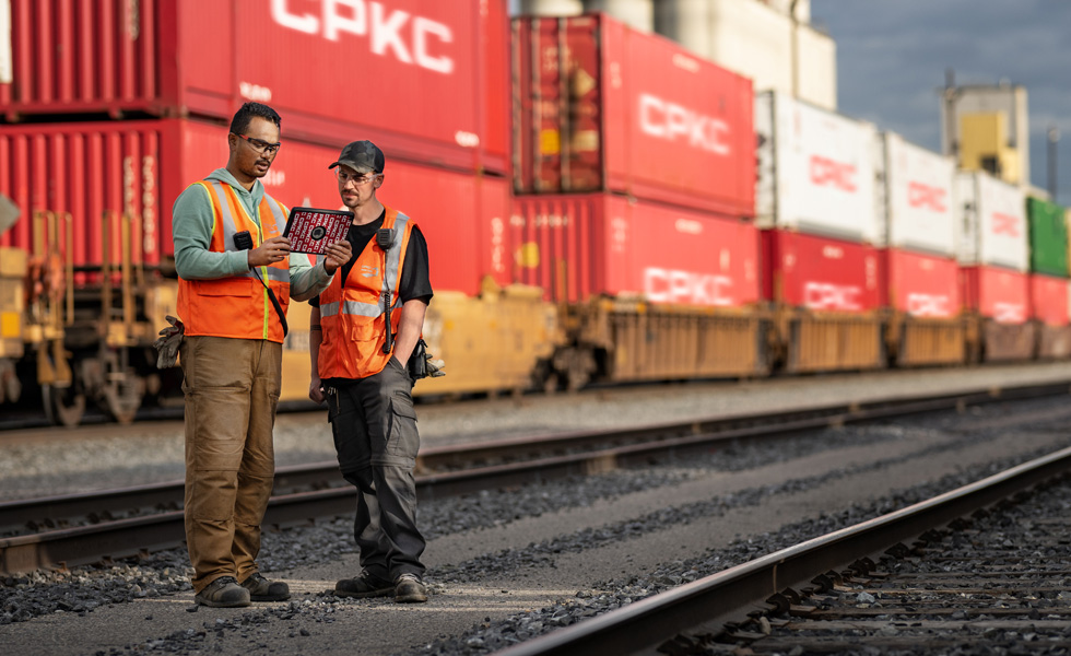 Zwei Bahnarbeiter blicken auf ein iPad, während sie auf einem Bahnhof neben einem stehenden Zug stehen, der Schiffscontainer transportiert