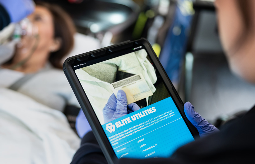 The Elite Utilities app is open on iPad, and a paramedic scans a patient's drivers license, while the patient lays on a transport bed