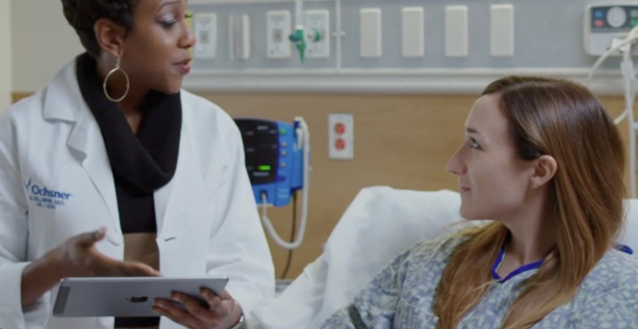 A provider with an iPad speaking to a patient in a hospital bed.