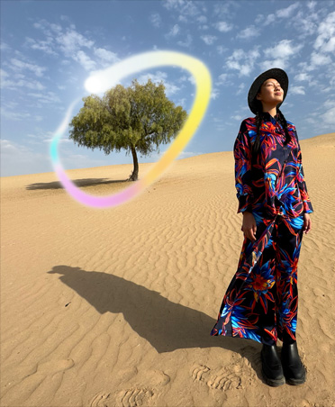 Picture of a woman in a multicolored dress standing in a desert landscape with a lone tree in the background circled in a glowing rainbow gradient showcasing Apple Intelligence capability to erase background objects