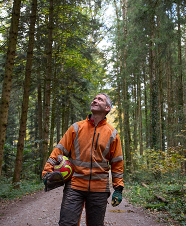 Man wearing an orange high-visibility jacket holding a hard hat while standing on a forest path in a sustainably managed forest in Switzerland